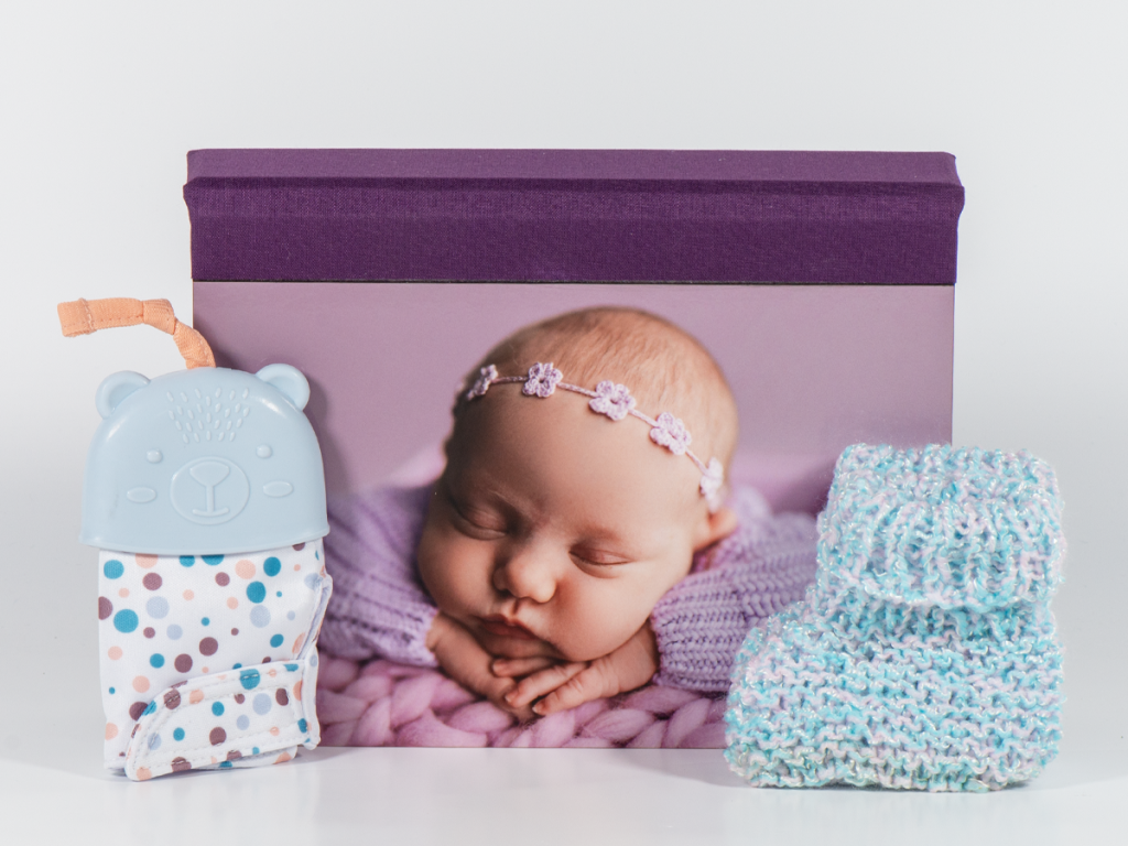 McKenna's Image Box featuring a newborn portrait printed on a ChromaLuxe cover paired with Plum Linen. Sitting infront of the box is a part of crocheted booties and a teether.