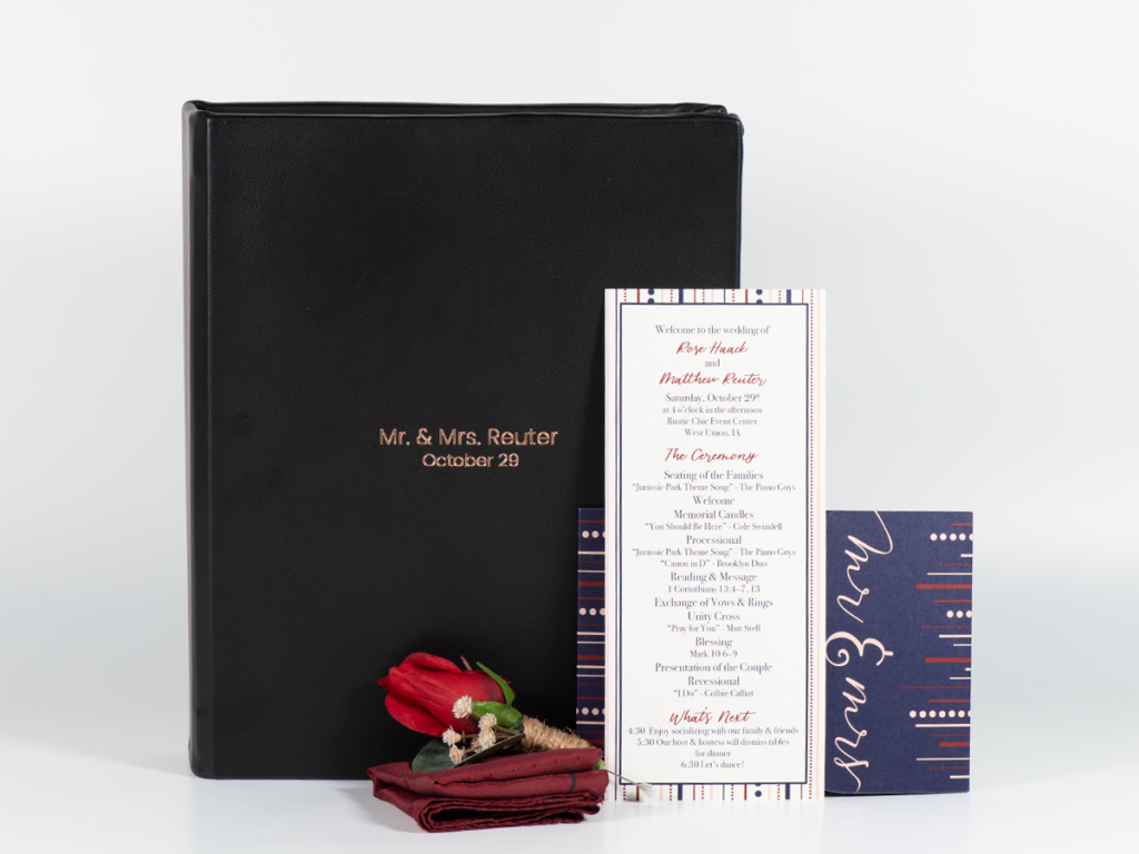 Black Leather Image Box with rose gold imprinting of first line "Mr. & Mrs. Reuter" and second line "October 29". Sitting propped against the Image Box is a wedding invite, wedding program, pocket square, and rose boutonniere.